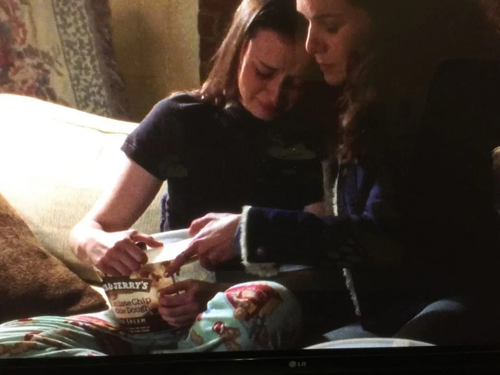 Two girls sitting on a couch together in their pajamas eating ice cream out of the tub. 